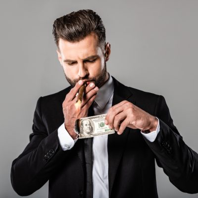 rich successful businessman in black suit lighting up cigar from burning dollar isolated on grey