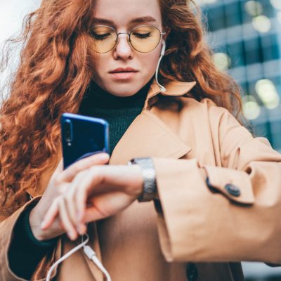 Office worker young woman wearing fashion clothes waits for business meeting looking at clock and holds laptop in arms standing against financial center.