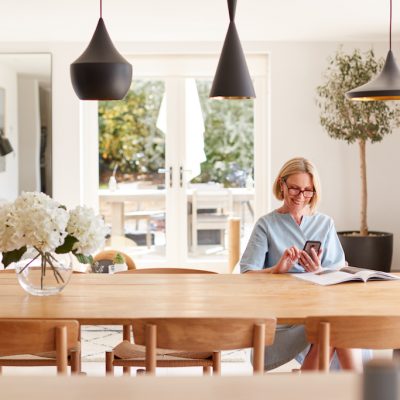 Senior Woman Relaxing With Magazine At Home Looking At Mobile Phone Sitting At Dining Room Table